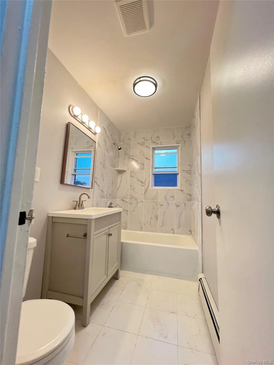Beautifully renovated bathroom featuring a chic grey vanity with brushed nickel hardware and a sophisticated marble-patterned tile tub surround. The bright, clean finishes create a spa-like atmosphere. Beautifully renovated bathroom featuring a chic grey vanity with brushed nickel hardware and a sophisticated marble-patterned tile tub surround. The bright, clean finishes create a spa-like atmosphere.