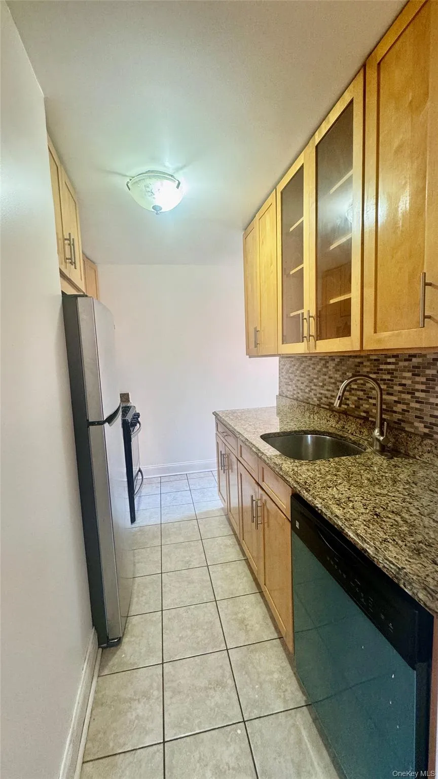 Kitchen featuring dishwashing machine, freestanding refrigerator, light tile patterned flooring, dark stone countertops, and backsplash Kitchen featuring dishwashing machine, freestanding refrigerator, light tile patterned flooring, dark stone countertops, and backsplash