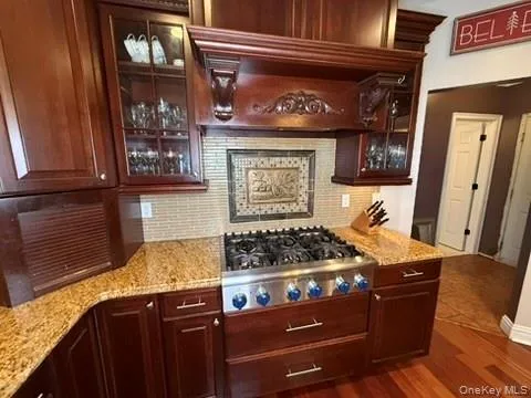 Kitchen with decorative backsplash, light stone counters, stainless steel gas stovetop, dark wood finished floors, and glass fronted cabinets Kitchen with decorative backsplash, light stone counters, stainless steel gas stovetop, dark wood finished floors, and glass fronted cabinets