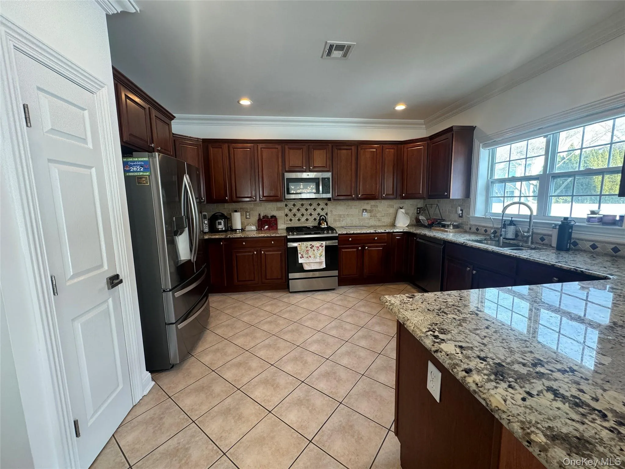 Kitchen featuring stainless steel appliances, crown molding, dark wood finish cabinetry, light stone countertops, and light tile patterned floors Kitchen featuring stainless steel appliances, crown molding, dark wood finish cabinetry, light stone countertops, and light tile patterned floors
