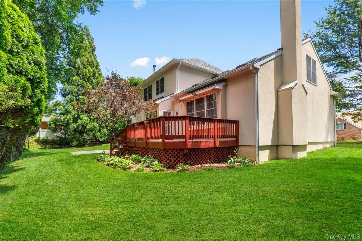 Rear exterior featuring an elevated wood deck with red railings, a stucco facade, a prominent chimney structure, and a large grassy yard Rear exterior featuring an elevated wood deck with red railings, a stucco facade, a prominent chimney structure, and a large grassy yard