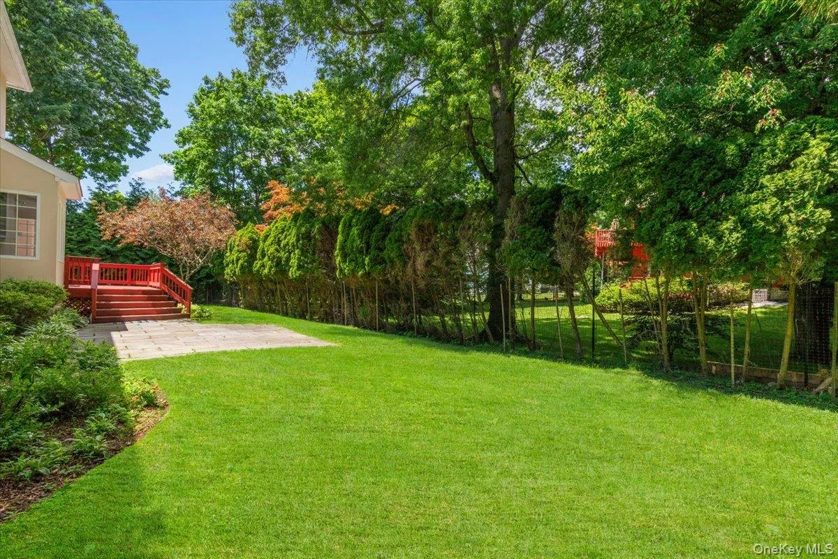 Expansive lawn area featuring a red-stained wood deck with stairs, a stone patio, and mature trees providing natural screening Expansive lawn area featuring a red-stained wood deck with stairs, a stone patio, and mature trees providing natural screening