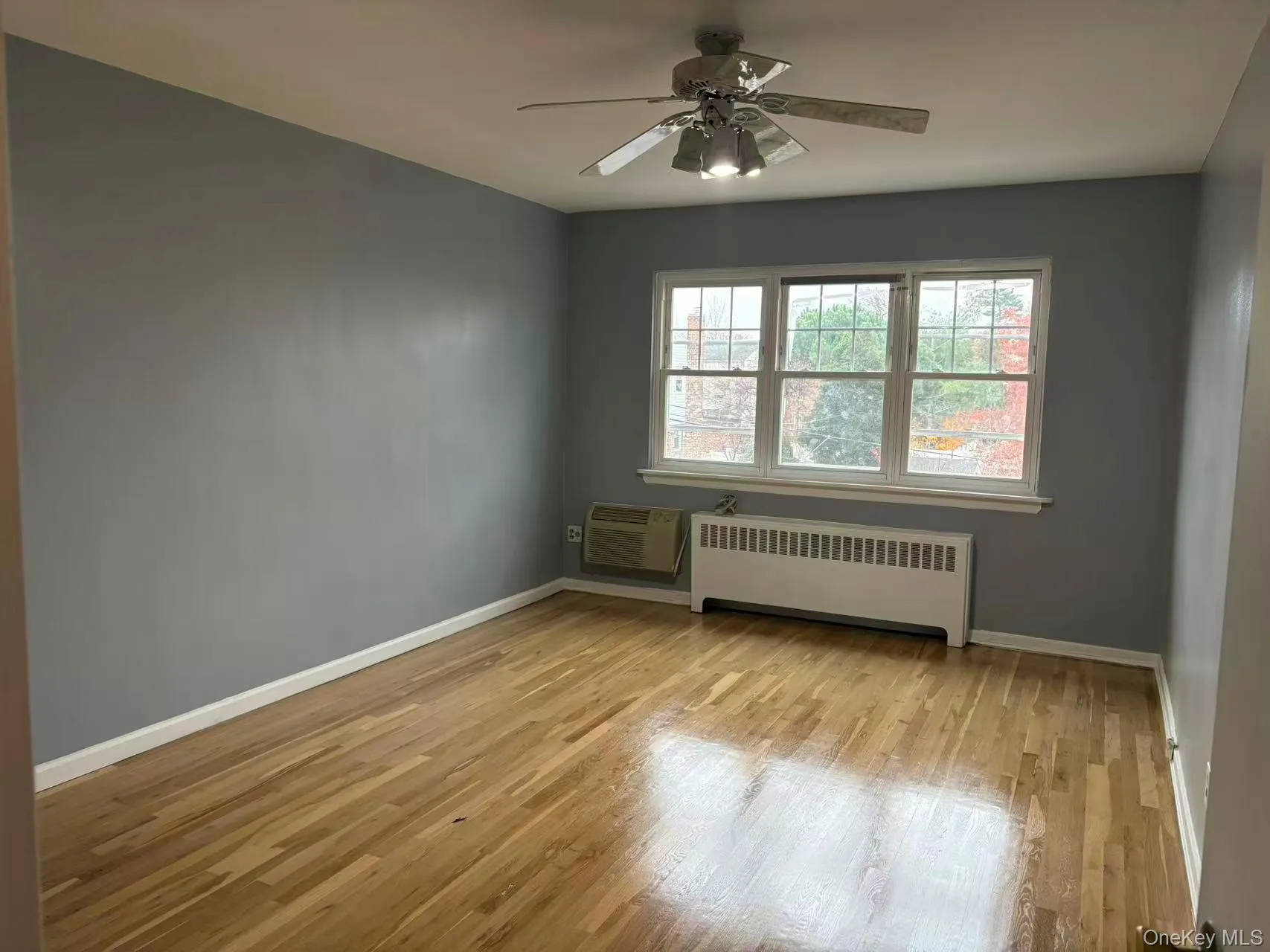 Empty room with radiator, light wood-type flooring, ceiling fan, and a wall mounted air conditioner Empty room with radiator, light wood-type flooring, ceiling fan, and a wall mounted air conditioner