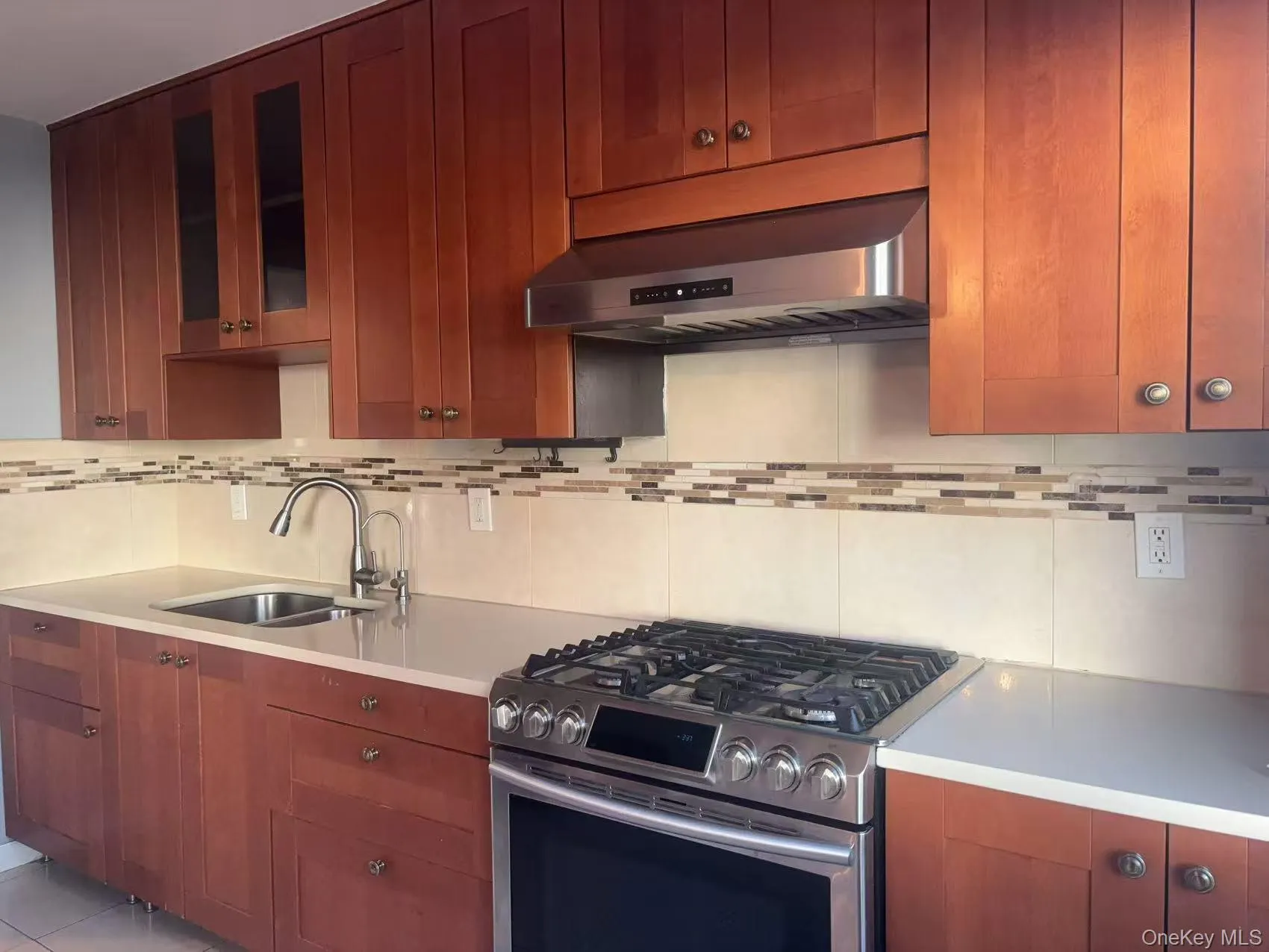 Kitchen featuring stainless steel range with gas cooktop, under cabinet range hood, glass insert cabinets, tasteful backsplash, and brown cabinetry Kitchen featuring stainless steel range with gas cooktop, under cabinet range hood, glass insert cabinets, tasteful backsplash, and brown cabinetry