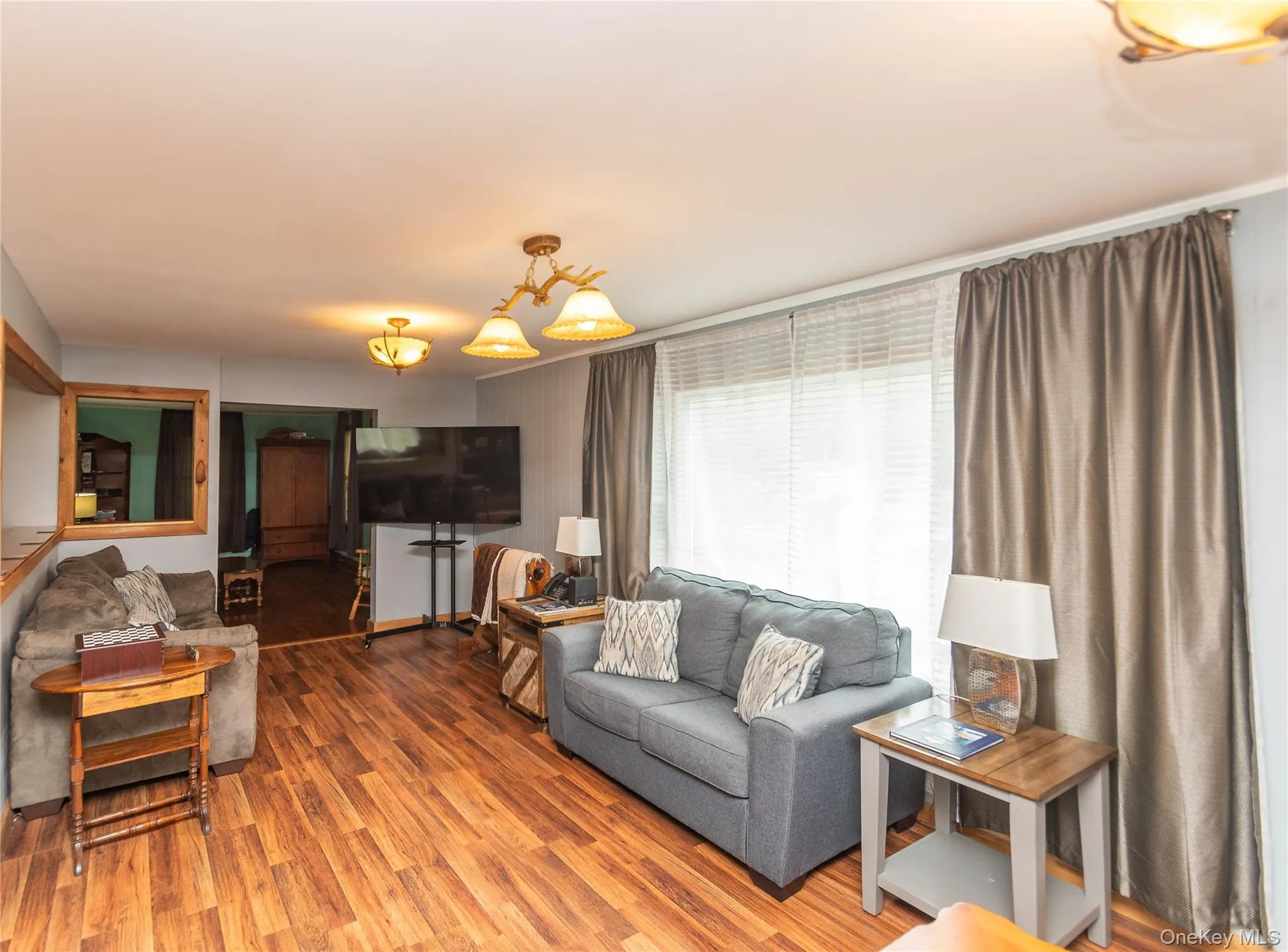 Living area featuring wood-finish flooring, two ceiling light fixtures, and a large window with blinds and drapes Living area featuring wood-finish flooring, two ceiling light fixtures, and a large window with blinds and drapes