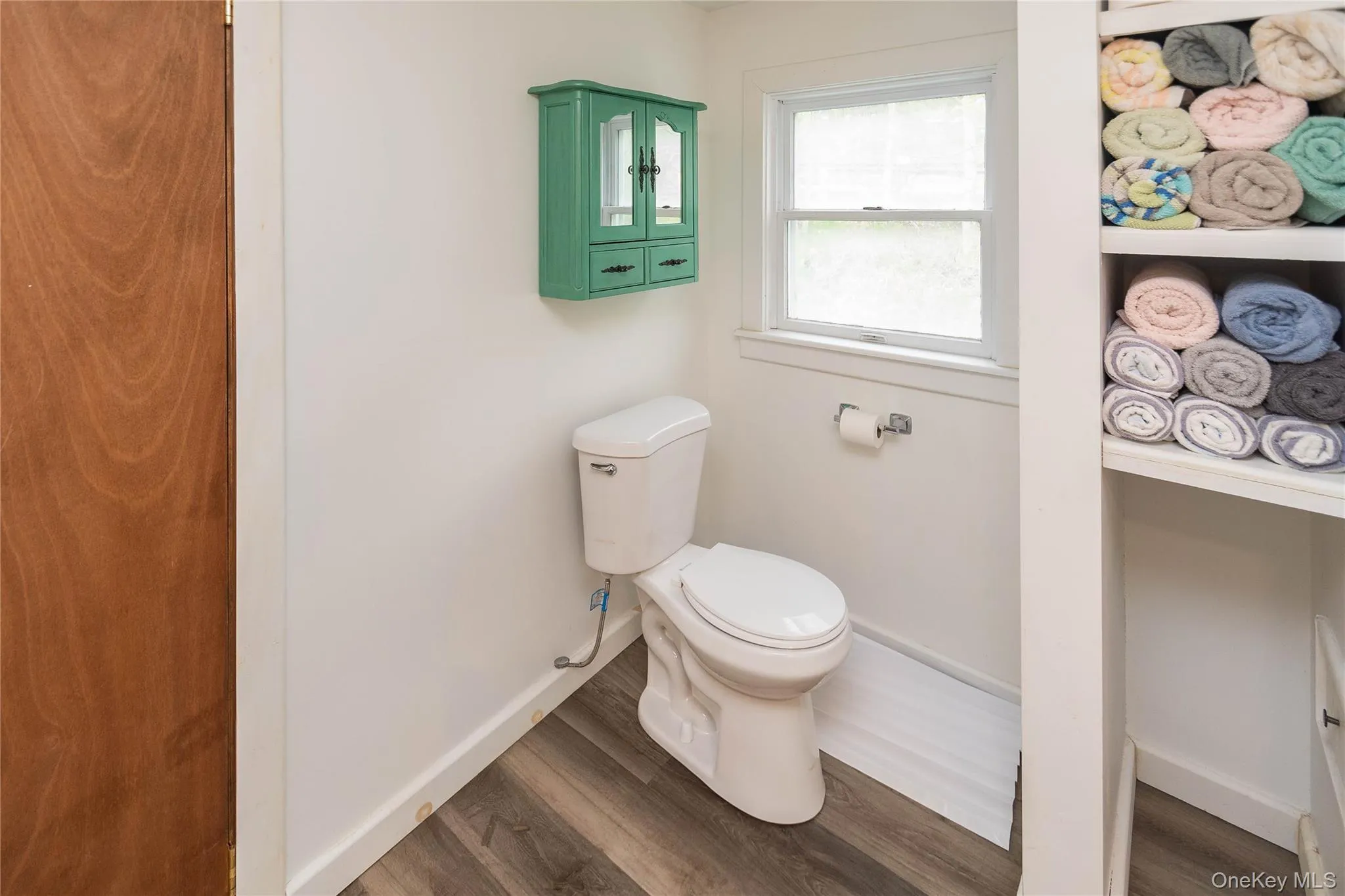 White toilet with wood-finish flooring, a bright window, and a wall-mounted green cabinet White toilet with wood-finish flooring, a bright window, and a wall-mounted green cabinet