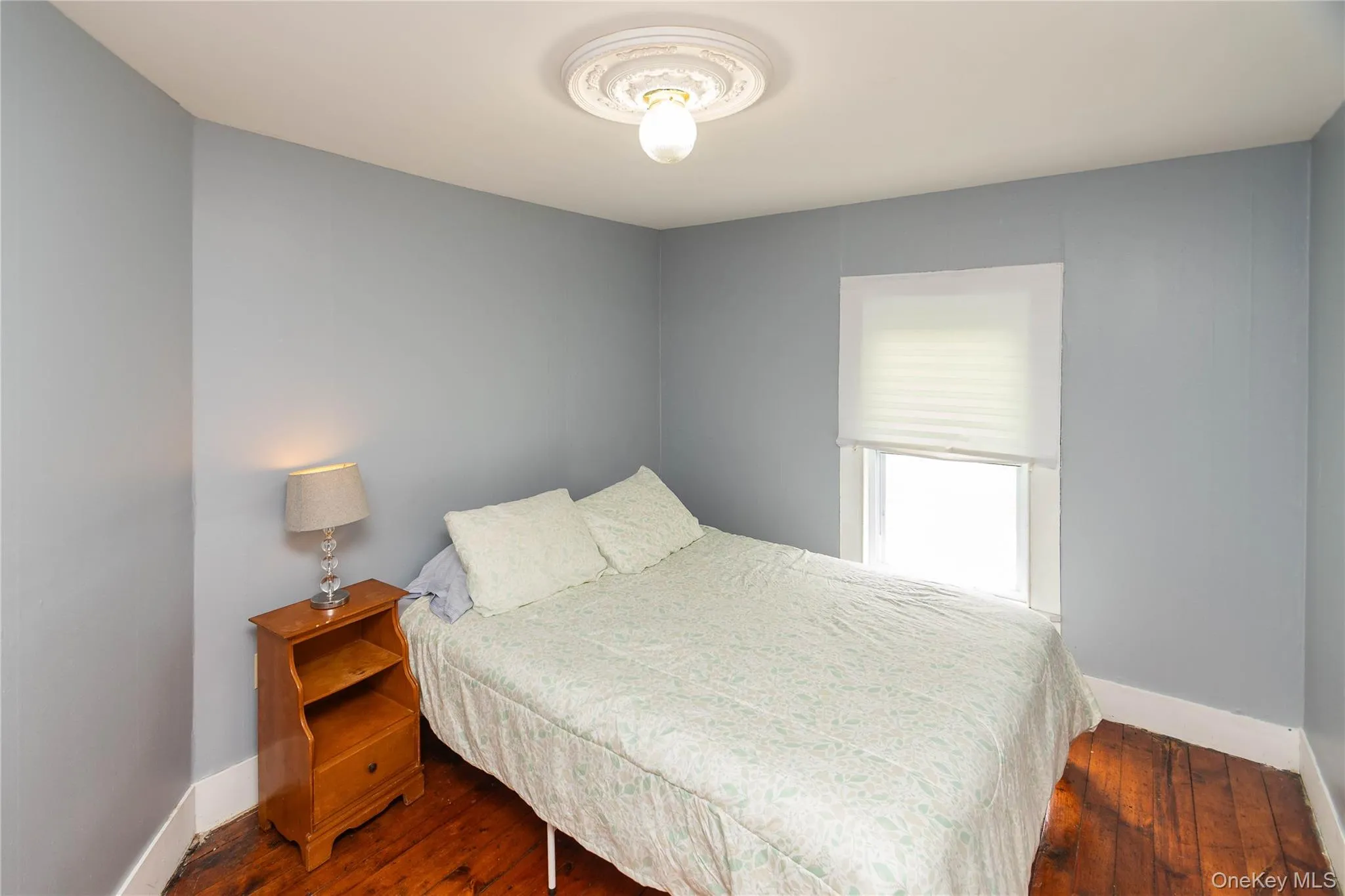 Bedroom featuring hardwood flooring, light blue wall paint, a ceiling medallion, a window with a cellular shade, and white trim Bedroom featuring hardwood flooring, light blue wall paint, a ceiling medallion, a window with a cellular shade, and white trim