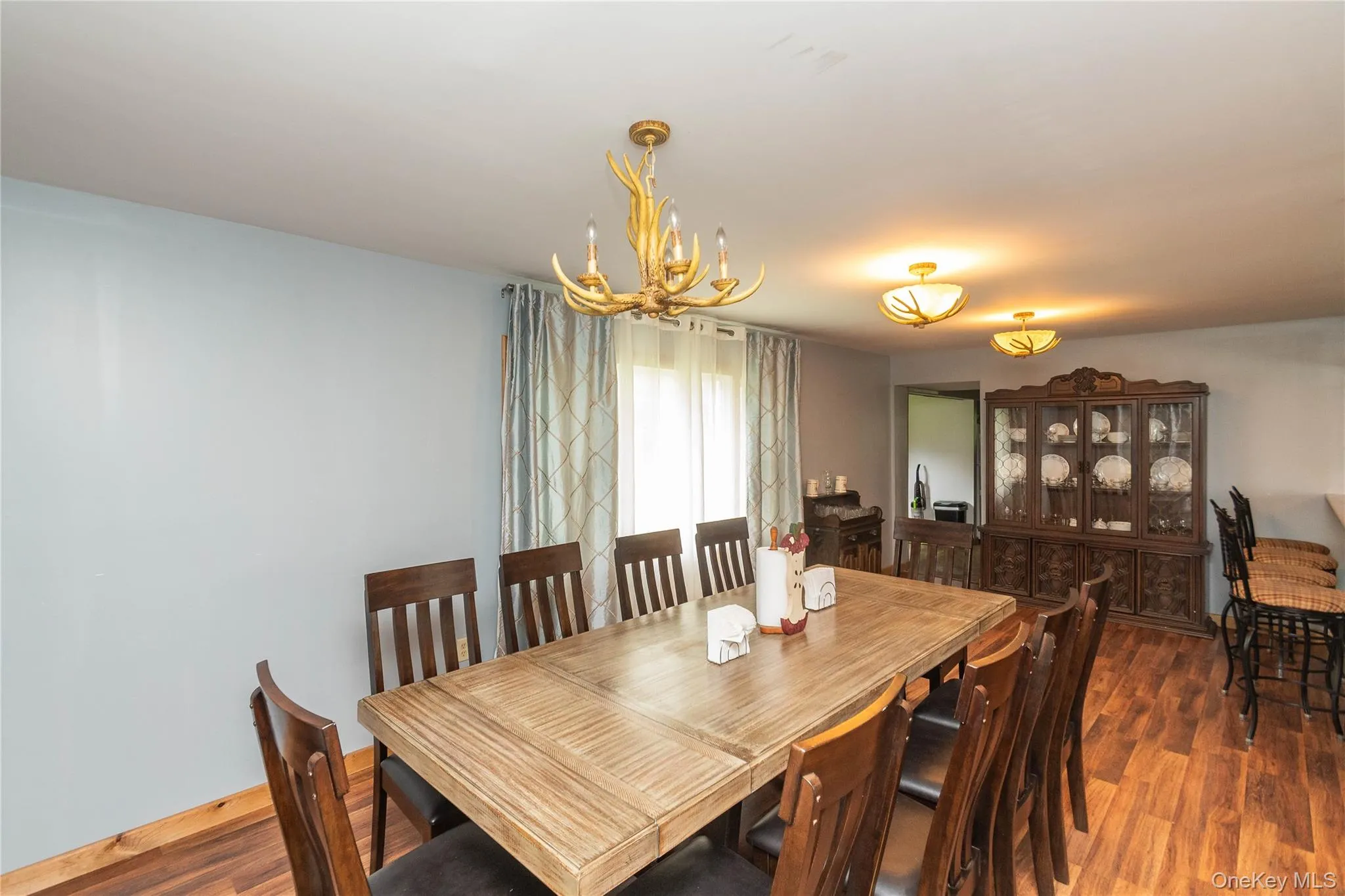 Spacious dining area featuring wood-finish flooring, light blue wall paint, and a window with patterned drapes Spacious dining area featuring wood-finish flooring, light blue wall paint, and a window with patterned drapes
