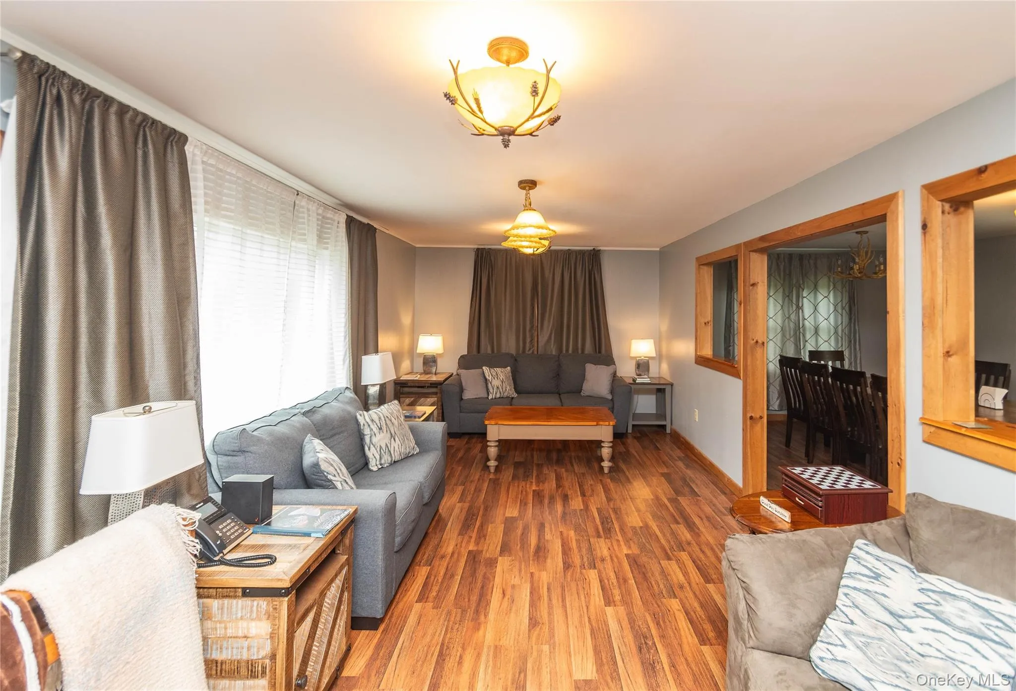 Expansive living area featuring wood-finish flooring, light gray wall tones, and two ornate ceiling light fixtures Expansive living area featuring wood-finish flooring, light gray wall tones, and two ornate ceiling light fixtures