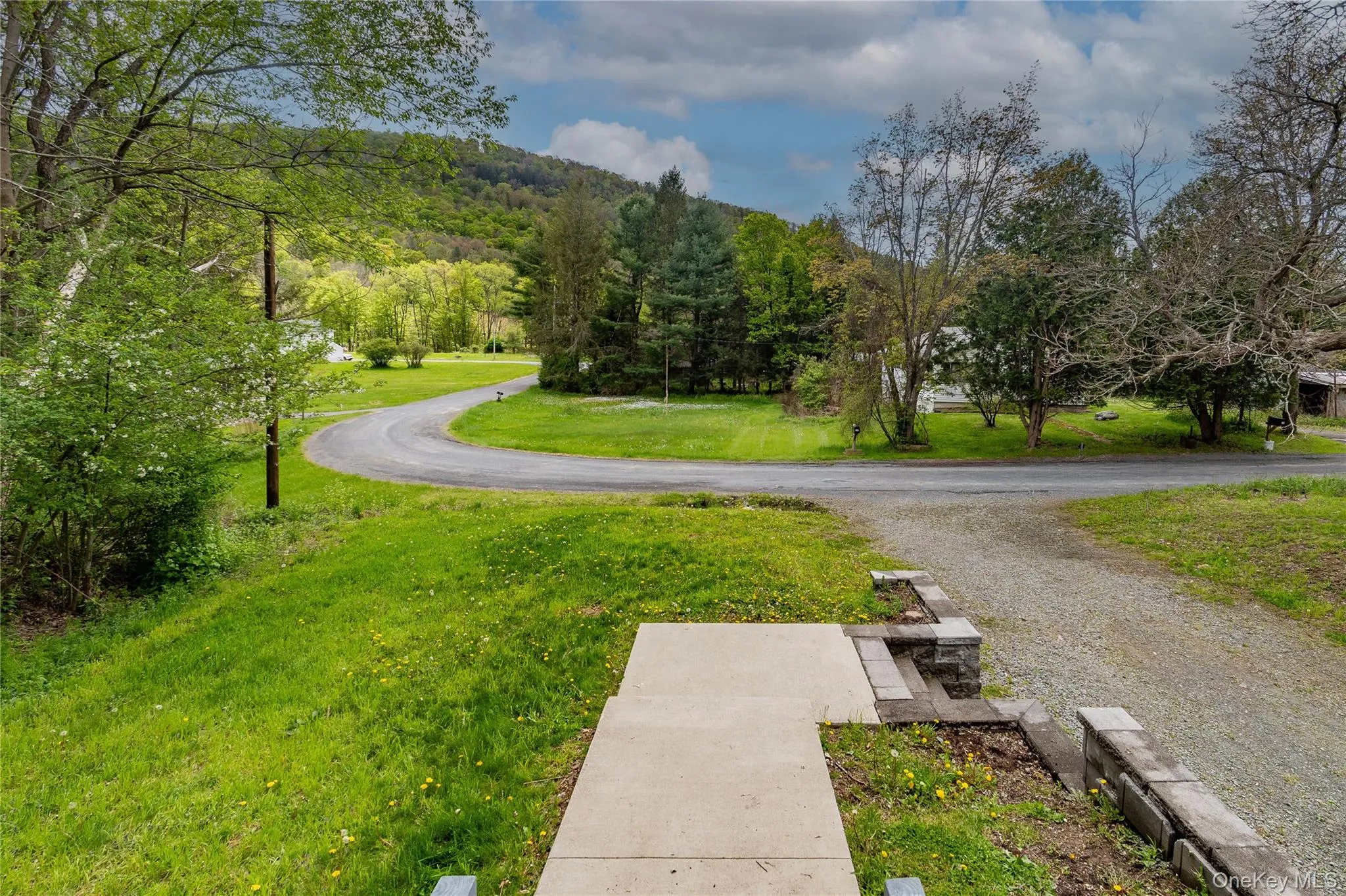 Expansive gravel driveway leading to a paved road, bordered by a concrete walkway and stacked stone retaining walls Expansive gravel driveway leading to a paved road, bordered by a concrete walkway and stacked stone retaining walls