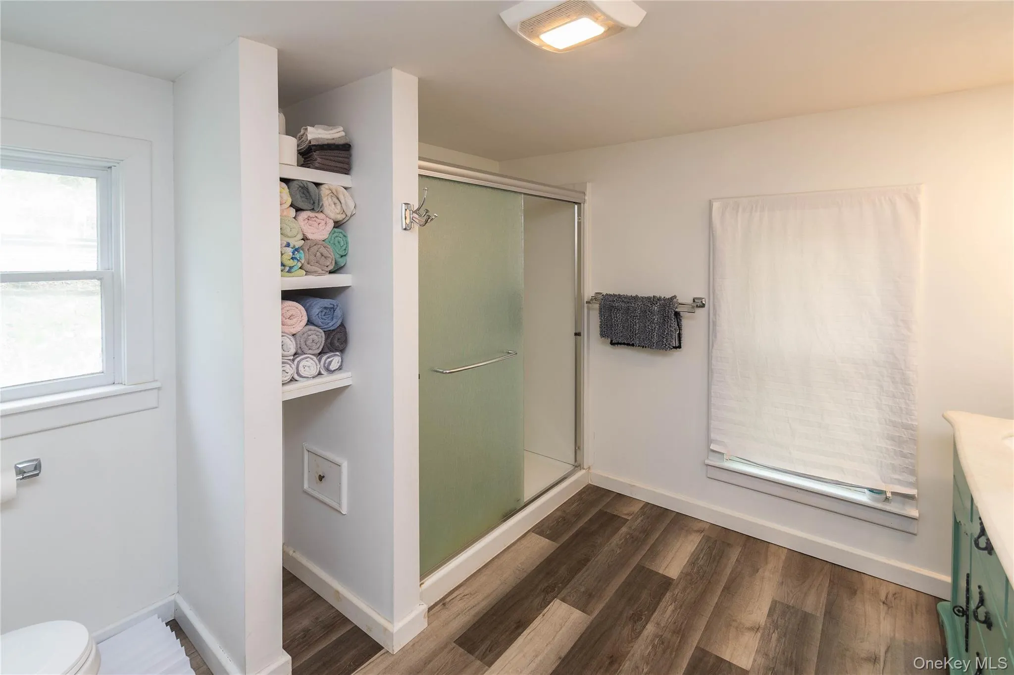 Walk-in shower with frosted glass enclosure, built-in shelving, wood-finish flooring, recessed lighting, and a window with privacy covering Walk-in shower with frosted glass enclosure, built-in shelving, wood-finish flooring, recessed lighting, and a window with privacy covering