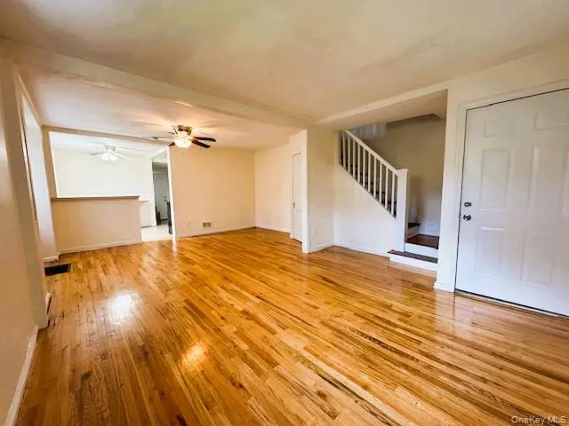 Spacious interior featuring wood-finish flooring, a white paneled entry door, a staircase with white balusters, a ceiling fan with light fixture, and neutral painted walls Spacious interior featuring wood-finish flooring, a white paneled entry door, a staircase with white balusters, a ceiling fan with light fixture, and neutral painted walls