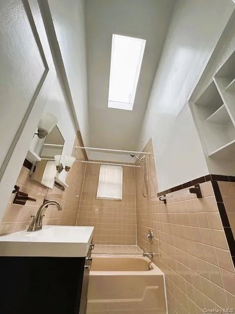 Bathroom featuring a rectangular Skylight, a built-in shower/tub combination, beige tiled walls with a dark accent border, a vanity with a white countertop, and integrated shelving Bathroom featuring a rectangular Skylight, a built-in shower/tub combination, beige tiled walls with a dark accent border, a vanity with a white countertop, and integrated shelving