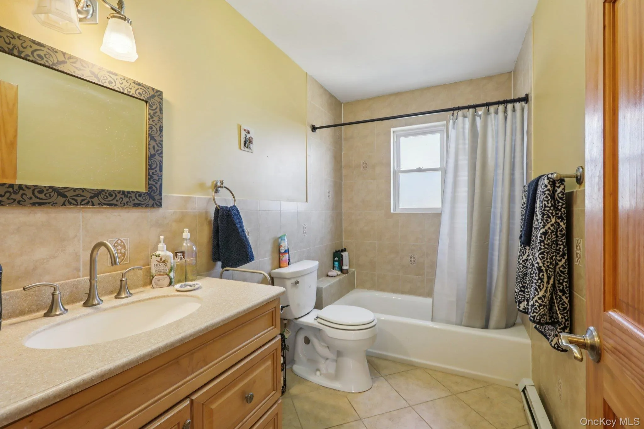 Bathroom with vanity, baseboard heating, tile walls, shower / bath combo with shower curtain, and light tile patterned flooring Bathroom with vanity, baseboard heating, tile walls, shower / bath combo with shower curtain, and light tile patterned flooring