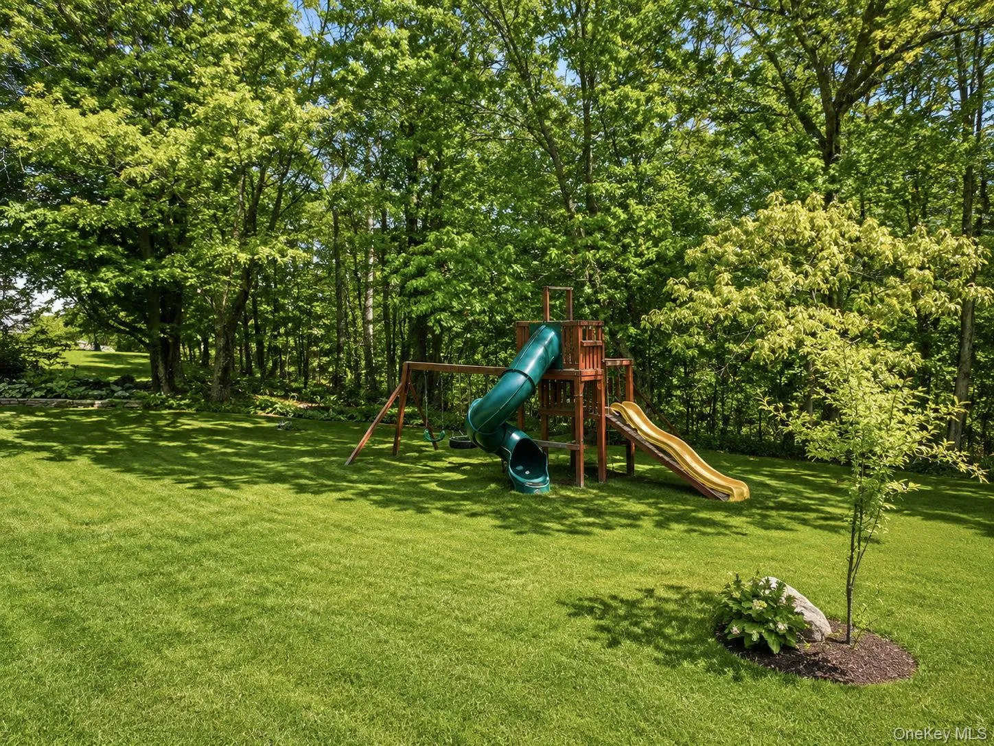 Expansive green lawn surrounded by mature trees, featuring a wooden playset with multiple slides and a swing component Expansive green lawn surrounded by mature trees, featuring a wooden playset with multiple slides and a swing component