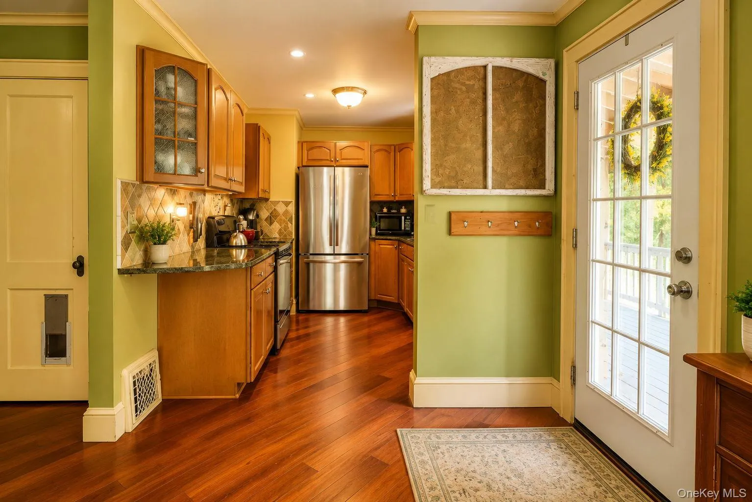 Kitchen entryway featuring Brazilian cherry wood flooring, light wood cabinetry, stainless steel appliances, a tiled backsplash, and a glass-paneled exterior door Kitchen entryway featuring Brazilian cherry wood flooring, light wood cabinetry, stainless steel appliances, a tiled backsplash, and a glass-paneled exterior door