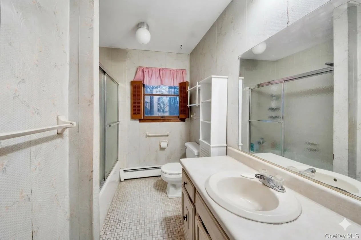 Bathroom featuring vanity, baseboard heating, bath / shower combo with glass door, light tile patterned floors, and wallpapered walls Bathroom featuring vanity, baseboard heating, bath / shower combo with glass door, light tile patterned floors, and wallpapered walls