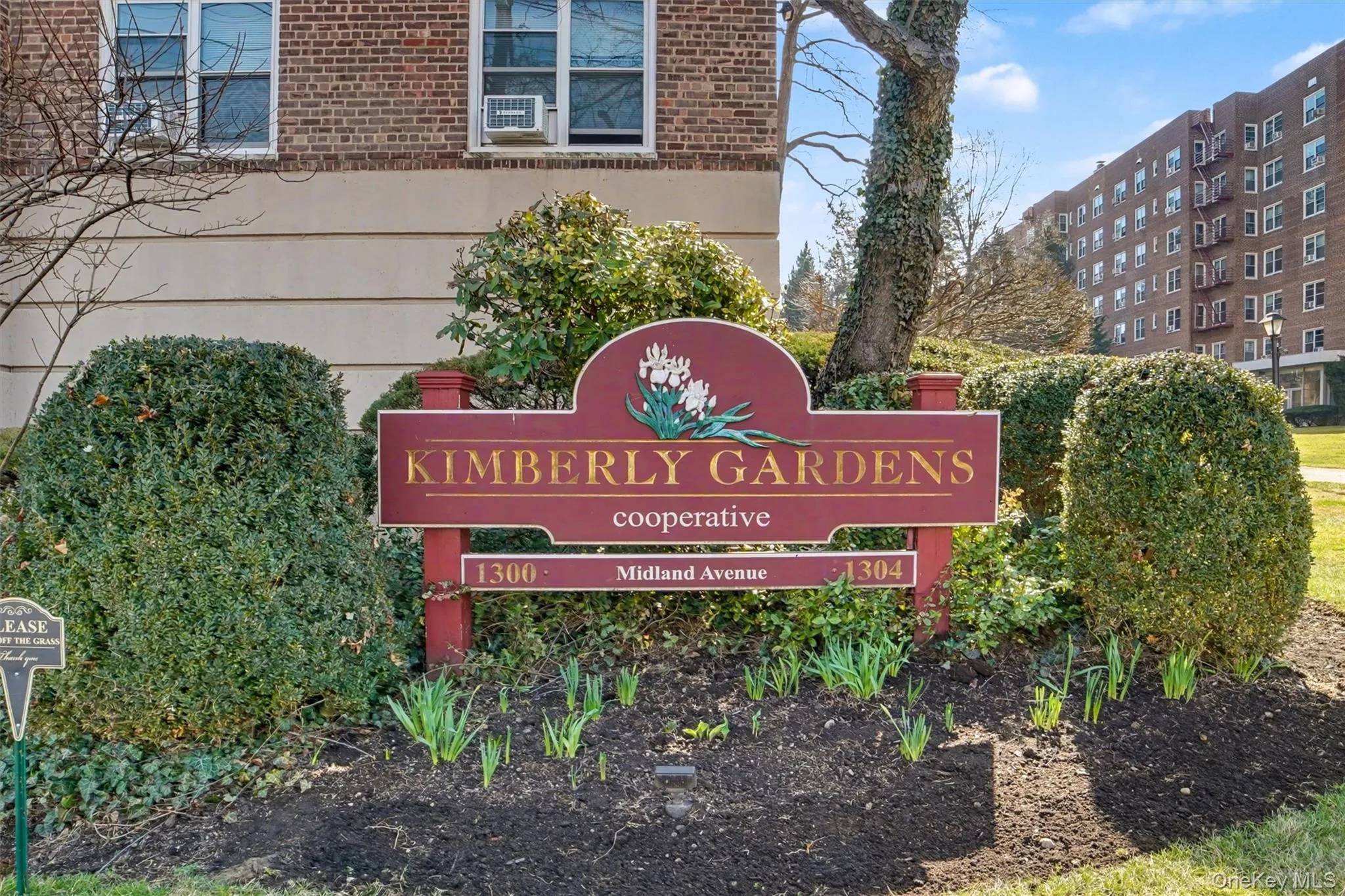 Kimberly Gardens cooperative entrance sign with manicured shrubbery, nascent plantings, and a partial view of a multi-story brick building Kimberly Gardens cooperative entrance sign with manicured shrubbery, nascent plantings, and a partial view of a multi-story brick building