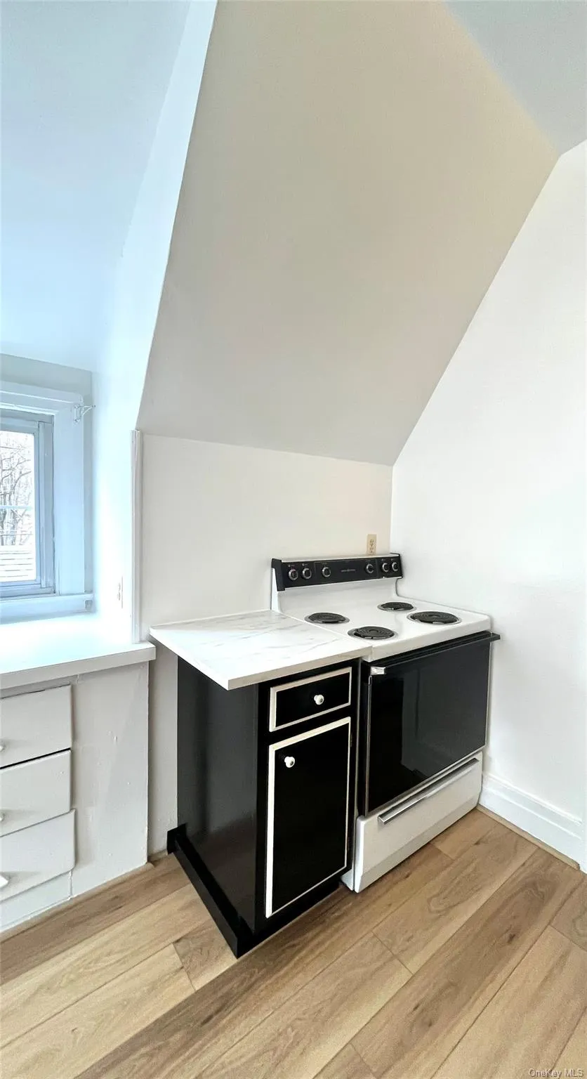 Area featuring white range with electric cooktop, vaulted ceiling, light wood-style floors, and dark cabinets Area featuring white range with electric cooktop, vaulted ceiling, light wood-style floors, and dark cabinets