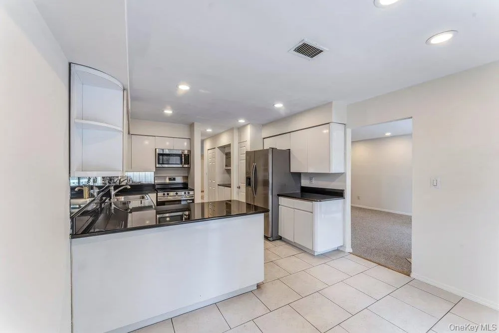 Kitchen with light carpet, a peninsula, stainless steel appliances, dark stone counters, and white cabinetry Kitchen with light carpet, a peninsula, stainless steel appliances, dark stone counters, and white cabinetry