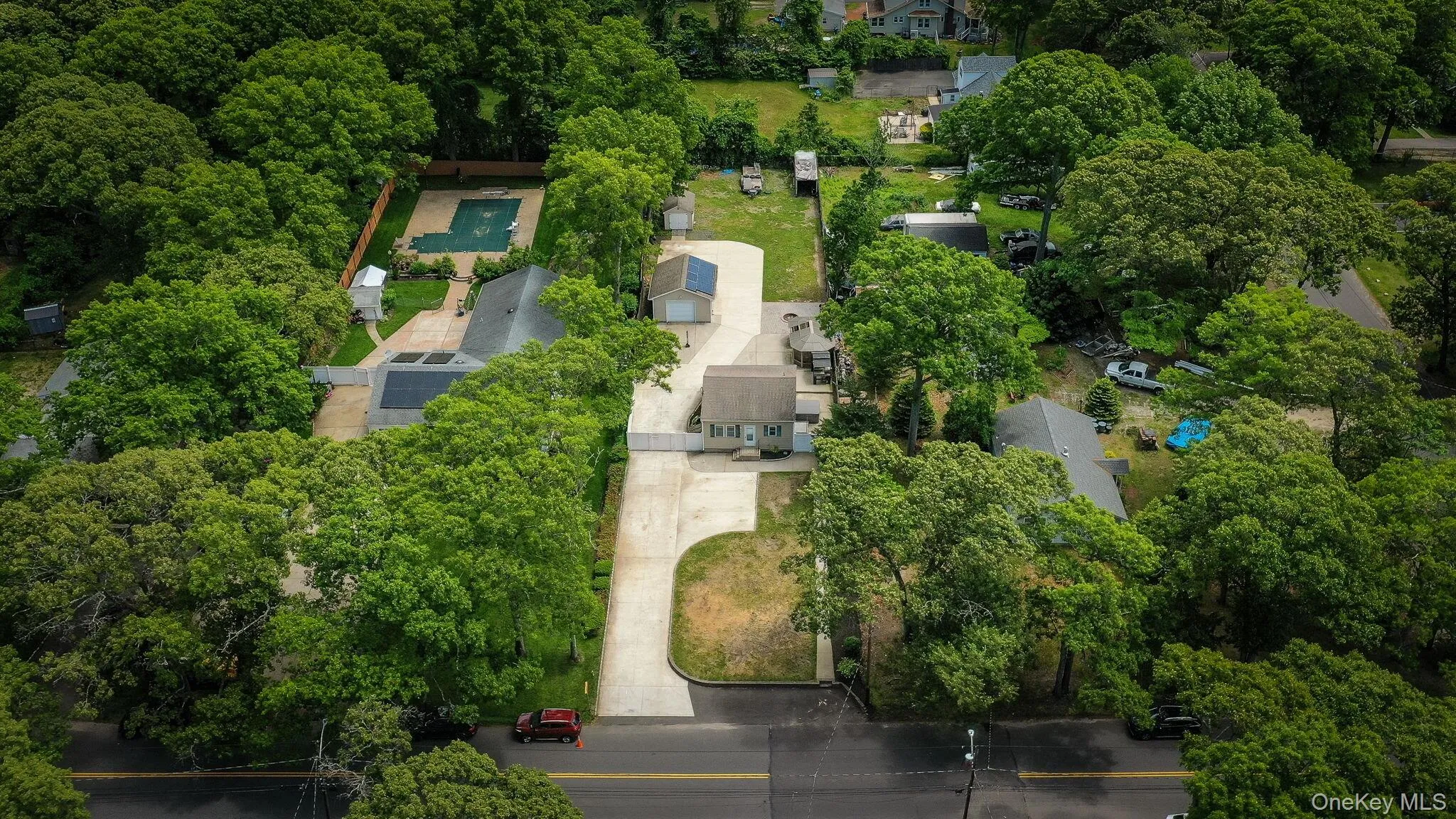 Aerial property view of expansive paved driveway leading to a single-story dwelling with a tan siding exterior, green shutters, white PVC fence Aerial property view of expansive paved driveway leading to a single-story dwelling with a tan siding exterior, green shutters, white PVC fence
