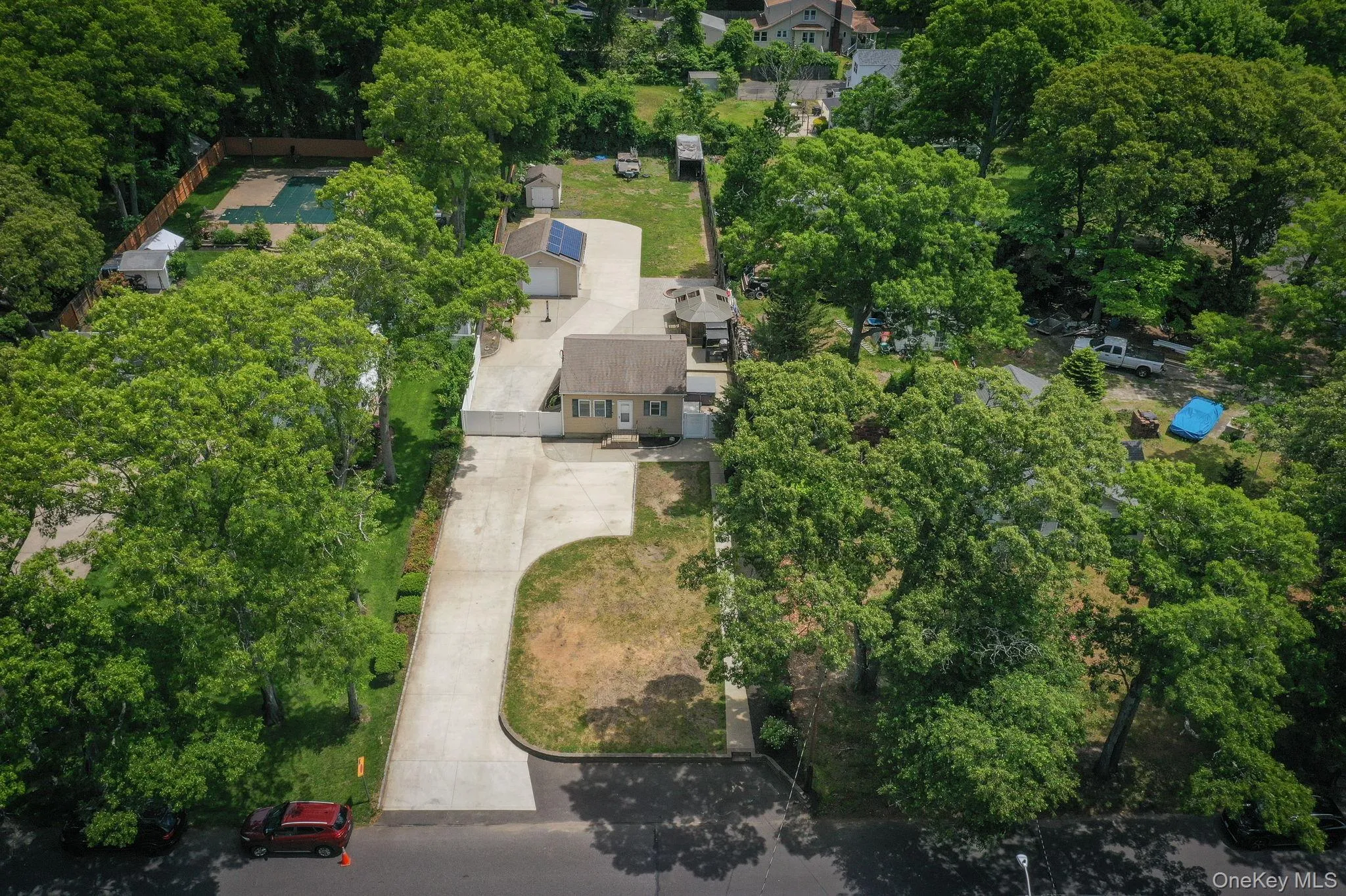 Aerial property view of expansive paved driveway leading to a single-story dwelling with a tan siding exterior, green shutters, white PVC fence Aerial property view of expansive paved driveway leading to a single-story dwelling with a tan siding exterior, green shutters, white PVC fence