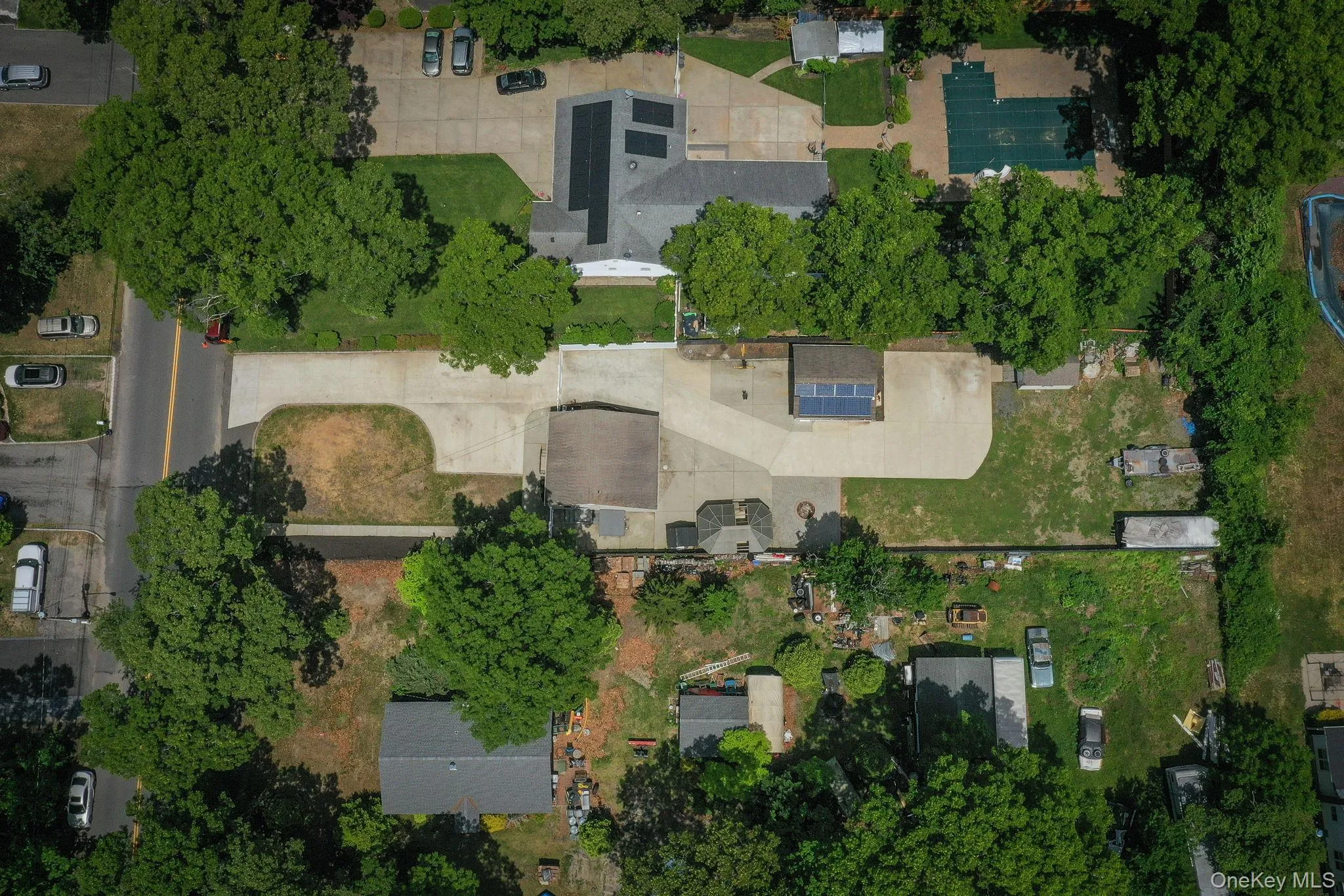 Aerial property view of expansive paved driveway leading to a single-story dwelling with a tan siding exterior, green shutters, white PVC fence Aerial property view of expansive paved driveway leading to a single-story dwelling with a tan siding exterior, green shutters, white PVC fence