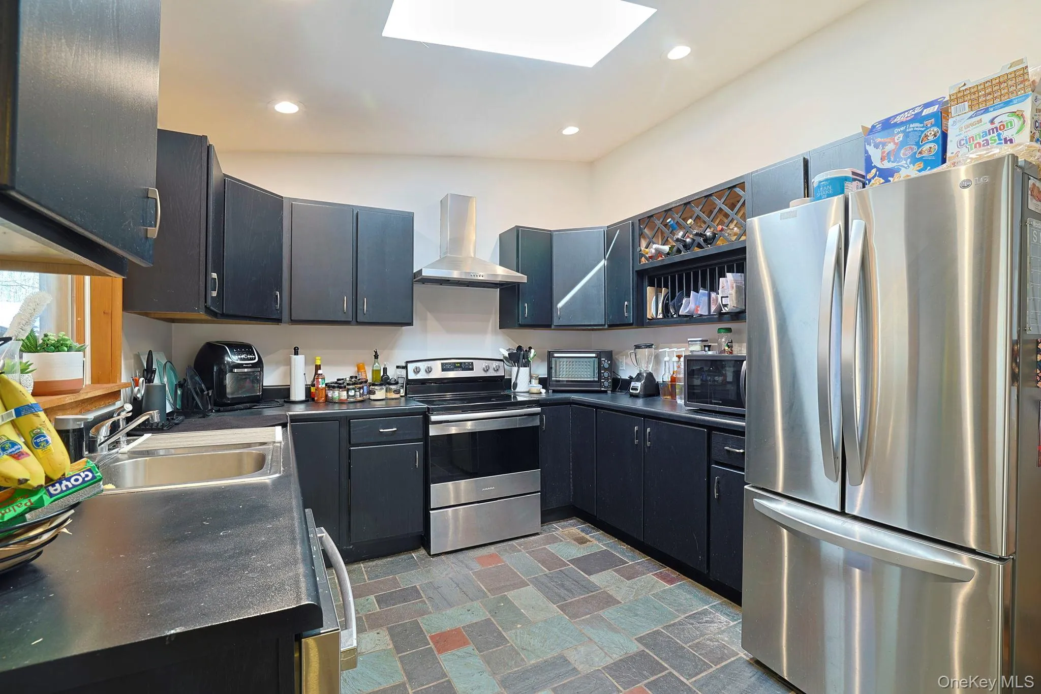 Kitchen featuring dark countertops, a sink, a skylight, wall chimney exhaust hood, and appliances with stainless steel finishes Kitchen featuring dark countertops, a sink, a skylight, wall chimney exhaust hood, and appliances with stainless steel finishes