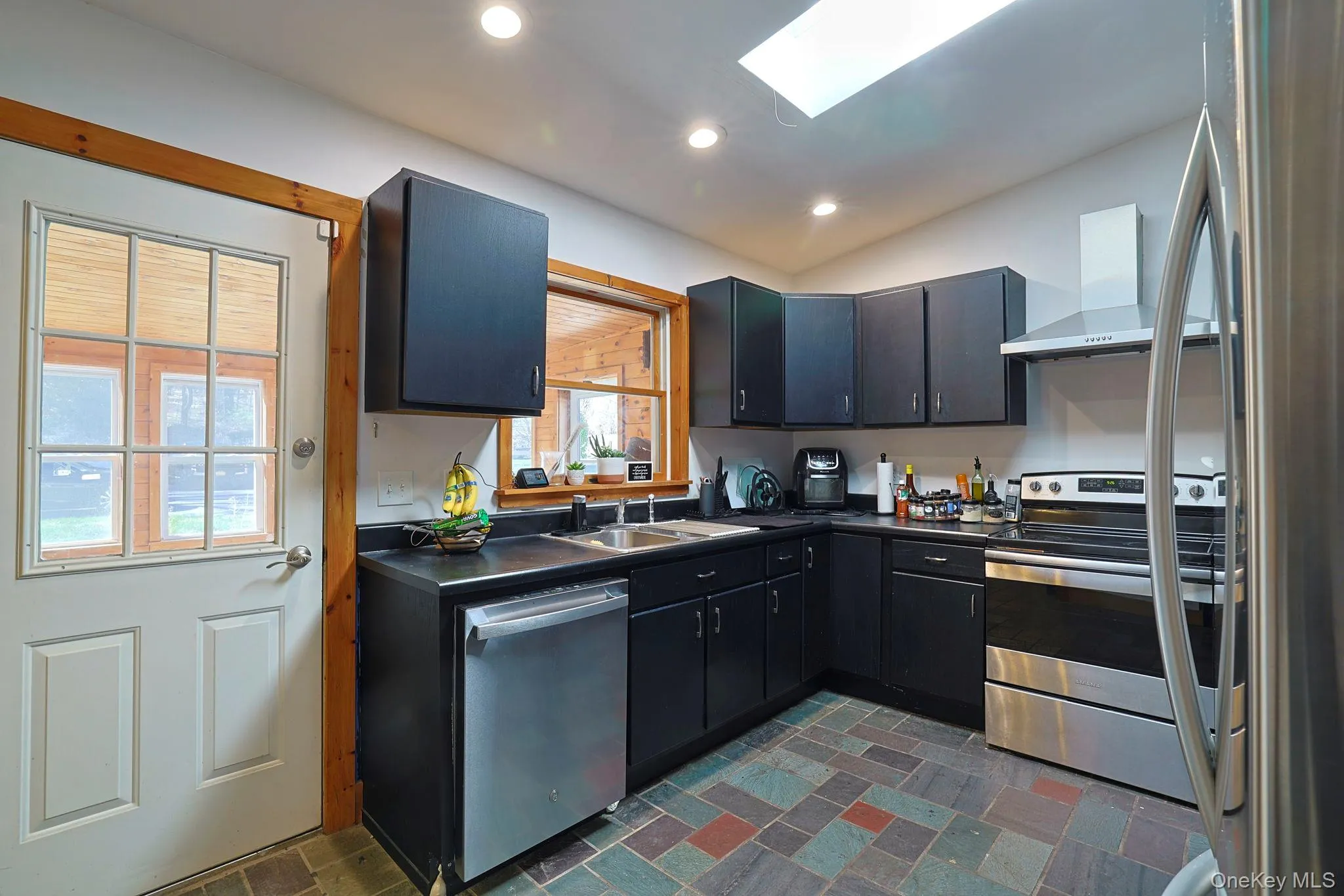 Kitchen with a sink, dark countertops, recessed lighting, wall chimney range hood, and appliances with stainless steel finishes Kitchen with a sink, dark countertops, recessed lighting, wall chimney range hood, and appliances with stainless steel finishes