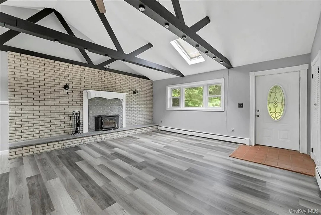 Unfurnished living room with a fireplace, lofted ceiling with skylight, hardwood / wood-style floors, brick wall, and a baseboard radiator Unfurnished living room with a fireplace, lofted ceiling with skylight, hardwood / wood-style floors, brick wall, and a baseboard radiator