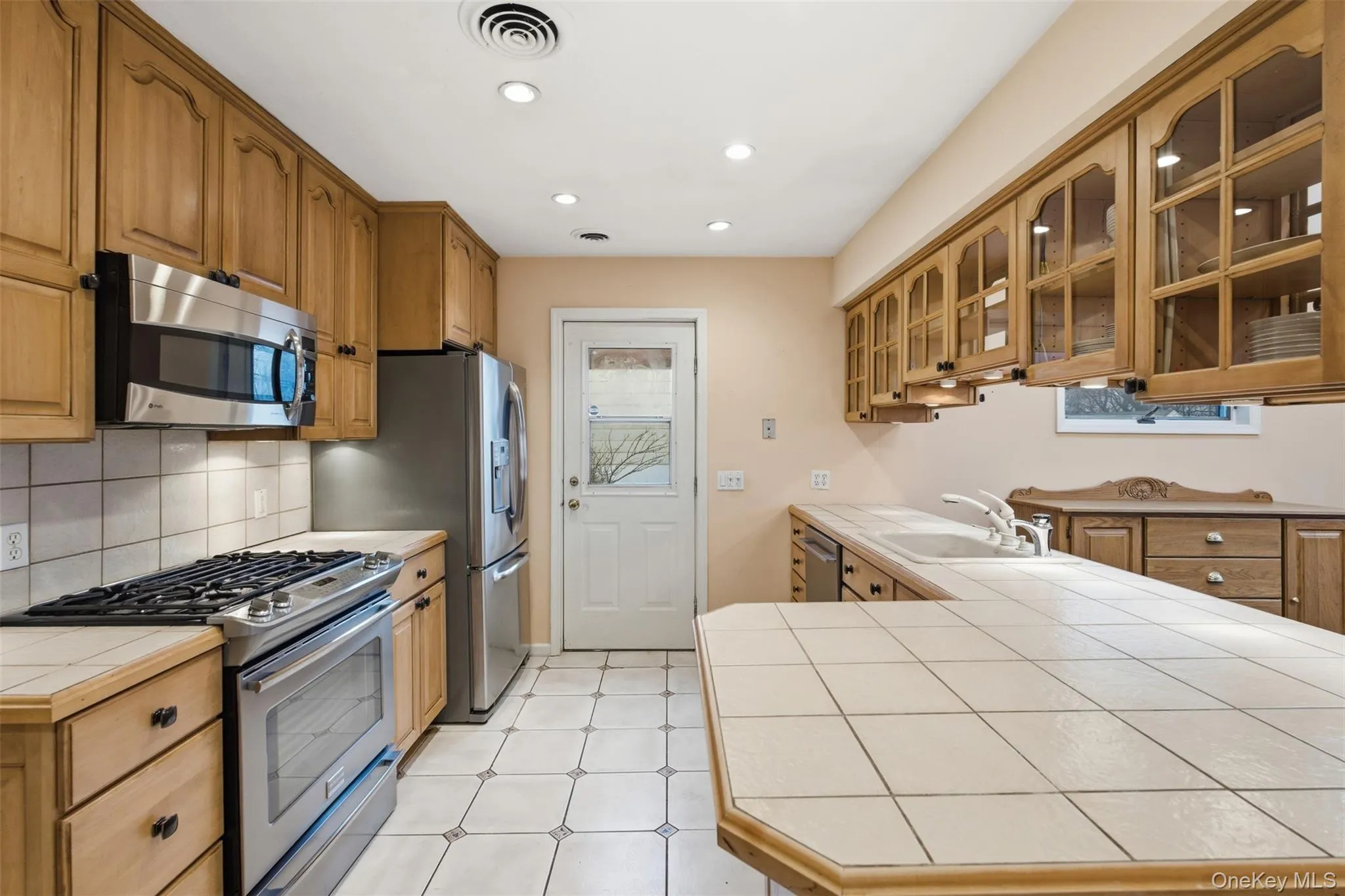 Kitchen with tile countertops, stainless steel appliances, glass fronted cabinets, wood finish cabinetry, and recessed lighting Kitchen with tile countertops, stainless steel appliances, glass fronted cabinets, wood finish cabinetry, and recessed lighting
