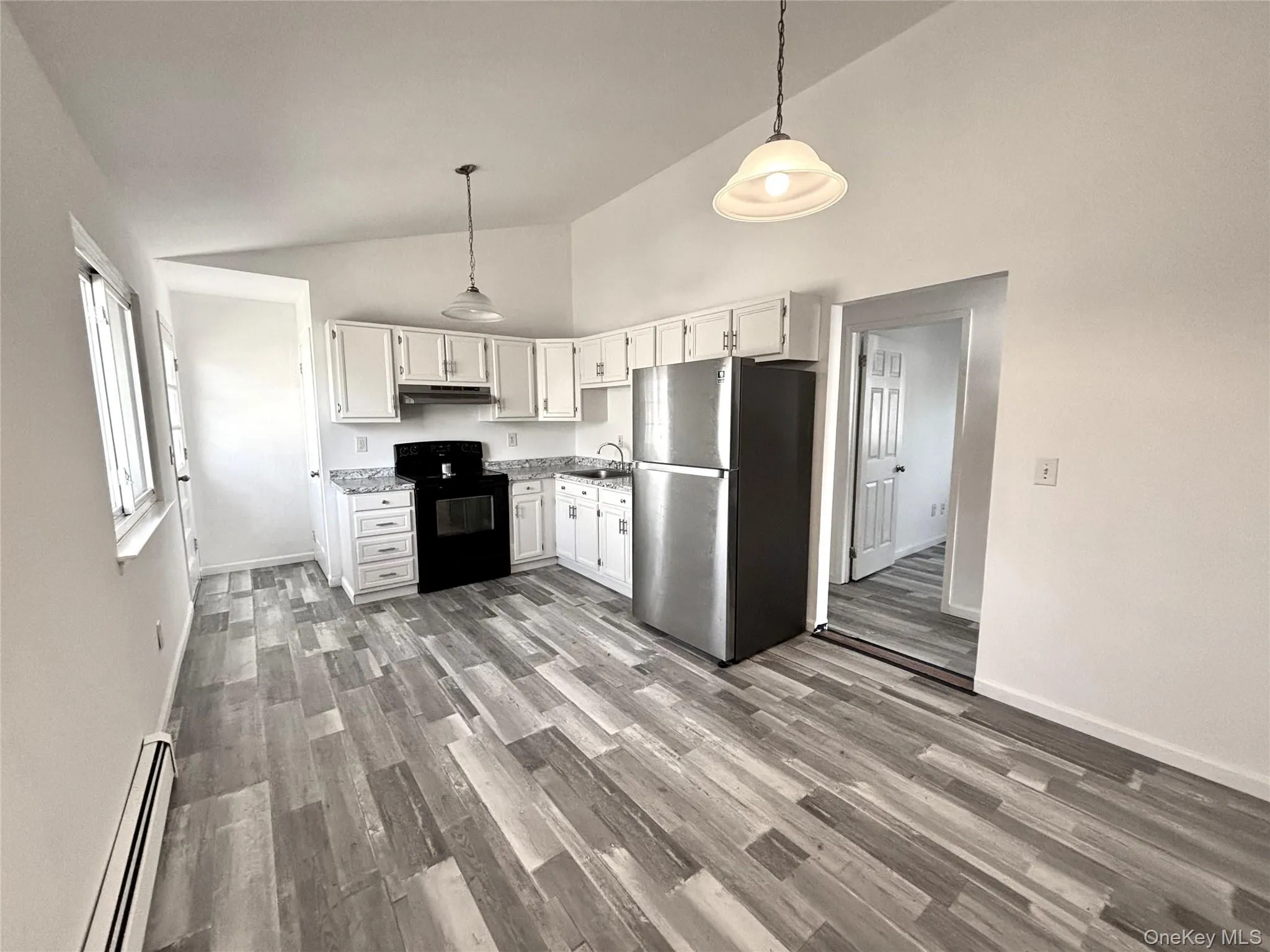 Kitchen with white cabinetry, vaulted ceiling, freestanding refrigerator, black electric range oven, and a baseboard, radiator and side entrance Kitchen with white cabinetry, vaulted ceiling, freestanding refrigerator, black electric range oven, and a baseboard, radiator and side entrance