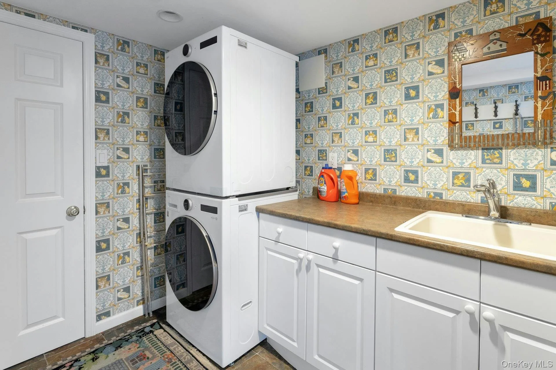 Washroom with wallpapered walls, stacked washer and clothes dryer, cabinet space, and dark tile patterned floors Washroom with wallpapered walls, stacked washer and clothes dryer, cabinet space, and dark tile patterned floors