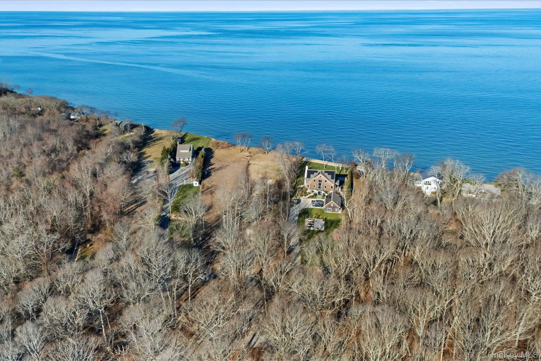 Aerial view showcasing the property's dramatic waterfront bluff setting, with open clearing near the shoreline, mature trees, and expansive open-water views beyond Aerial view showcasing the property's dramatic waterfront bluff setting, with open clearing near the shoreline, mature trees, and expansive open-water views beyond