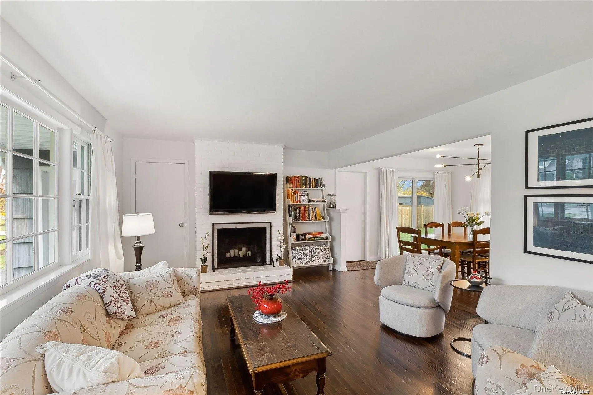 Living area featuring dark wood-finish flooring, a white brick fireplace, multiple windows with white trim, and a clear sightline into a dining area with a modern chandelier Living area featuring dark wood-finish flooring, a white brick fireplace, multiple windows with white trim, and a clear sightline into a dining area with a modern chandelier