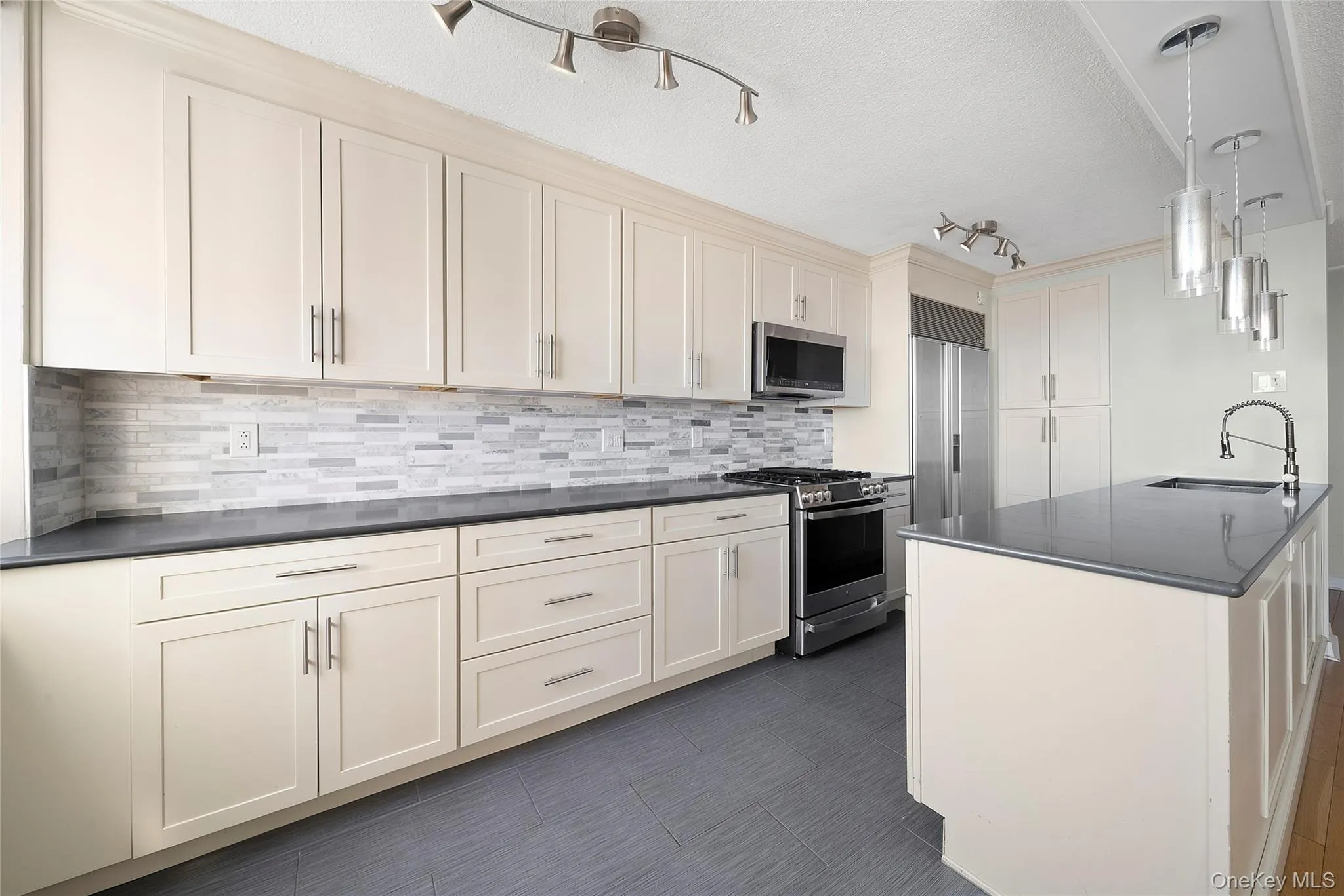 Kitchen featuring stainless steel appliances, hanging light fixtures, a textured ceiling, decorative backsplash, and cream cabinetry Kitchen featuring stainless steel appliances, hanging light fixtures, a textured ceiling, decorative backsplash, and cream cabinetry
