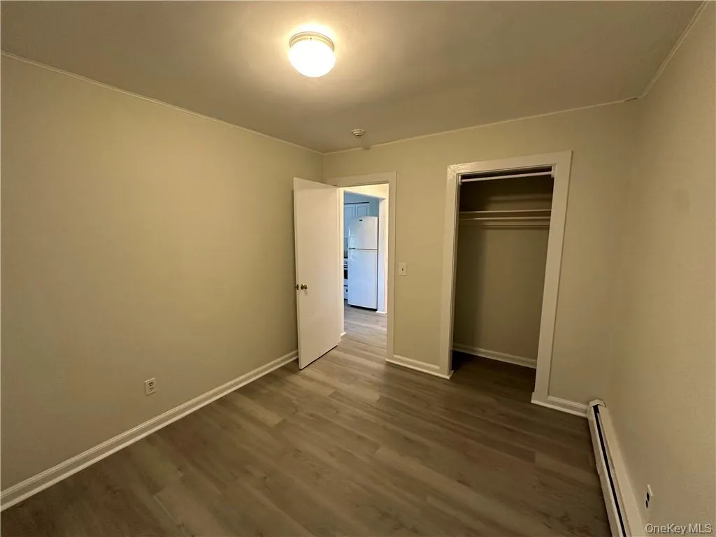 Unfurnished bedroom with a closet, baseboard heating, white refrigerator, and dark hardwood / wood-style flooring Unfurnished bedroom with a closet, baseboard heating, white refrigerator, and dark hardwood / wood-style flooring