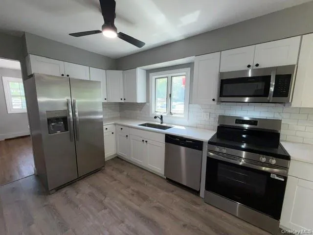 Kitchen featuring wood-finish flooring, white shaker cabinetry with dark hardware, stainless steel appliances, white subway tile backsplash, and light countertops Kitchen featuring wood-finish flooring, white shaker cabinetry with dark hardware, stainless steel appliances, white subway tile backsplash, and light countertops