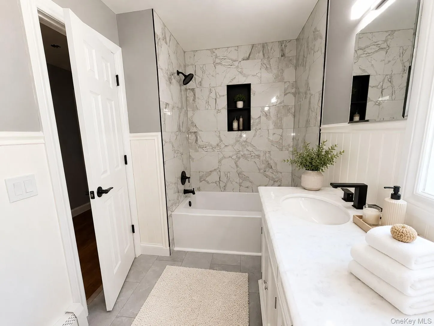 Bathroom featuring a white bathtub with a tile surround, a built-in shower niche, a white vanity with an integrated sink, a matte black faucet, and wainscoting Bathroom featuring a white bathtub with a tile surround, a built-in shower niche, a white vanity with an integrated sink, a matte black faucet, and wainscoting