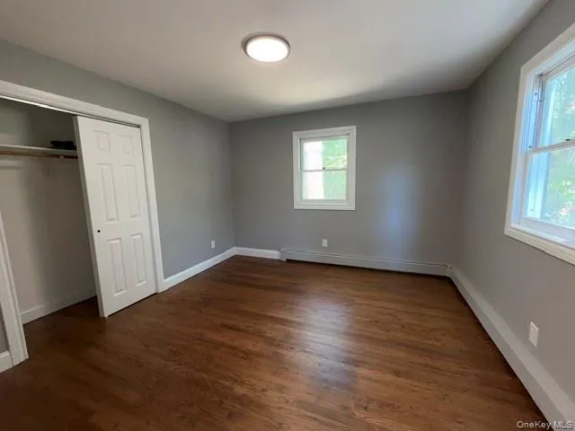 Bedroom featuring dark wood-finish flooring, two large windows, a flush-mount ceiling light, a built-in closet with sliding panel doors, and neutral grey walls Bedroom featuring dark wood-finish flooring, two large windows, a flush-mount ceiling light, a built-in closet with sliding panel doors, and neutral grey walls