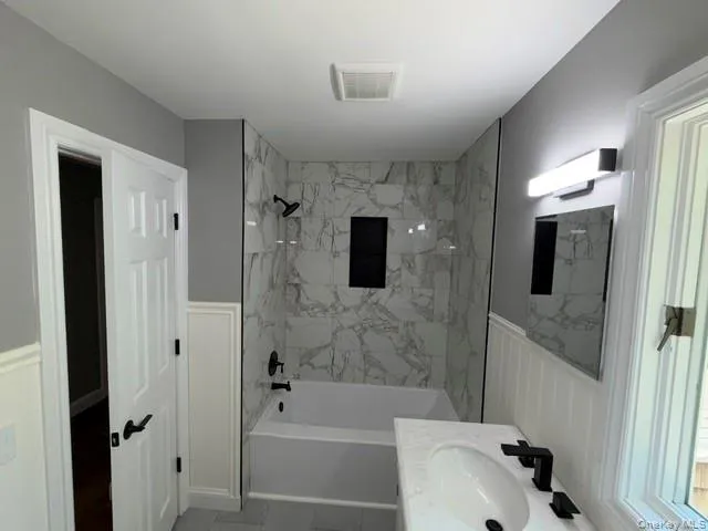 Bathroom featuring white wainscoting, a tiled tub and shower combination with a black niche, and a vanity with a white countertop and oval under-mount sink Bathroom featuring white wainscoting, a tiled tub and shower combination with a black niche, and a vanity with a white countertop and oval under-mount sink