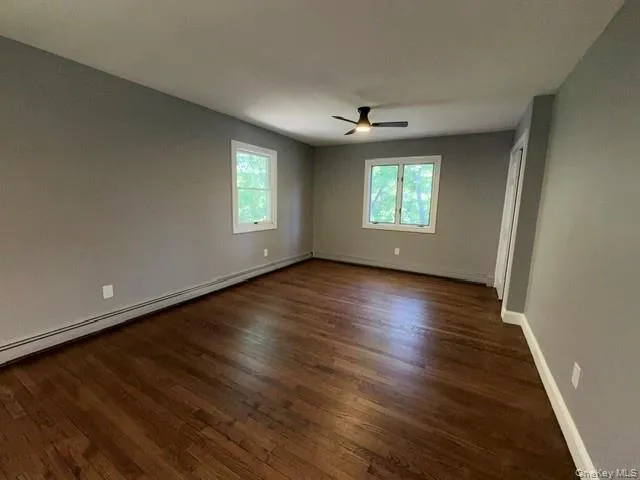 Spacious room featuring rich wood-finish flooring, two large windows, a ceiling fan with integrated lighting, and neutral wall tones Spacious room featuring rich wood-finish flooring, two large windows, a ceiling fan with integrated lighting, and neutral wall tones
