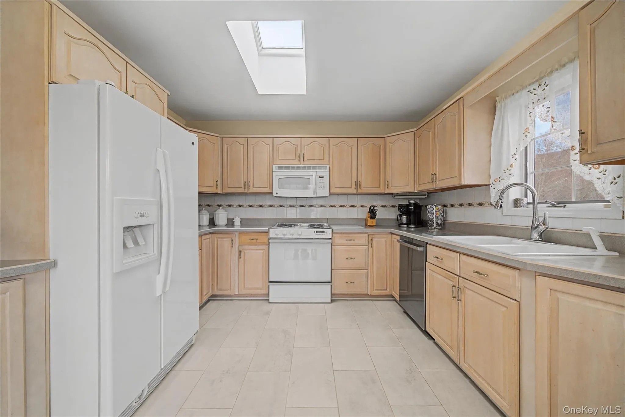 Kitchen with light wood finish cabinetry, white appliances, a skylight, light countertops, and decorative backsplash Kitchen with light wood finish cabinetry, white appliances, a skylight, light countertops, and decorative backsplash
