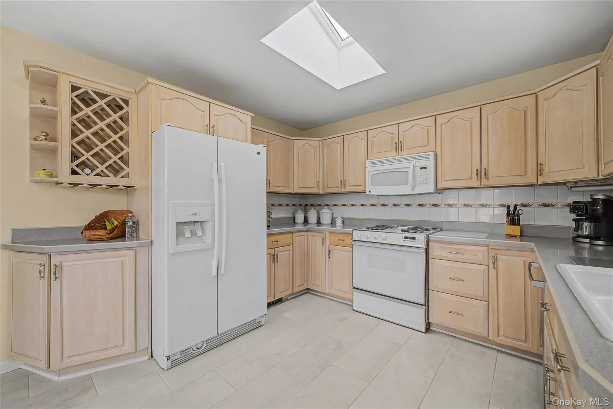 Kitchen featuring light wood finish cabinetry, white appliances, tasteful backsplash, light countertops, and a skylight Kitchen featuring light wood finish cabinetry, white appliances, tasteful backsplash, light countertops, and a skylight