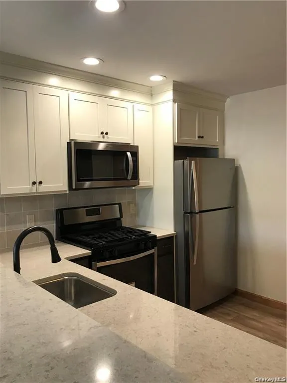 Kitchen featuring stainless steel appliances, tasteful backsplash, white cabinetry, light wood-style flooring, and light stone countertops Kitchen featuring stainless steel appliances, tasteful backsplash, white cabinetry, light wood-style flooring, and light stone countertops