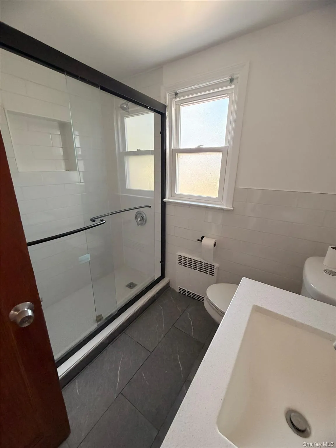 Full bath featuring tile walls, a shower stall, radiator heating unit, and a wainscoted wall Full bath featuring tile walls, a shower stall, radiator heating unit, and a wainscoted wall