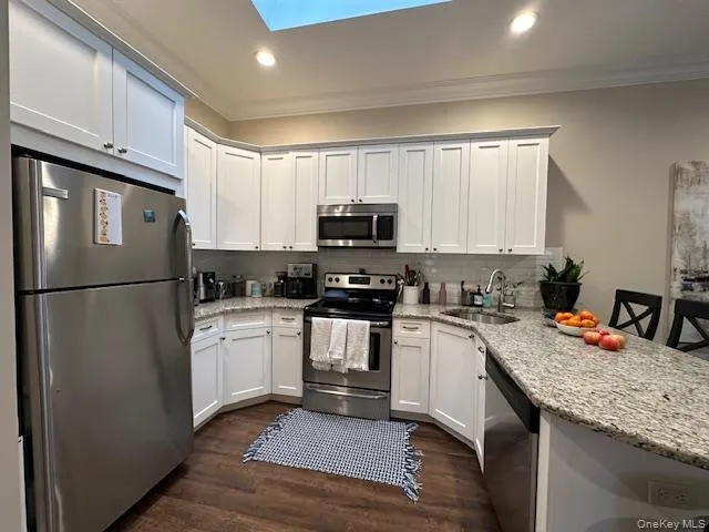 Kitchen with appliances with stainless steel finishes, ornamental molding, a peninsula, white cabinets, and dark wood-style flooring Kitchen with appliances with stainless steel finishes, ornamental molding, a peninsula, white cabinets, and dark wood-style flooring