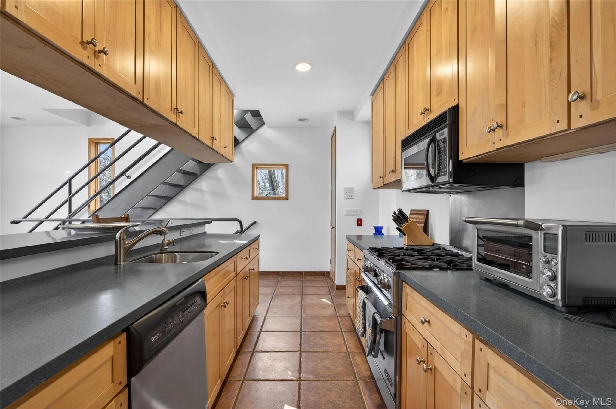 Galley kitchen featuring light wood cabinetry, dark countertops, a stainless steel sink, and tile flooring Galley kitchen featuring light wood cabinetry, dark countertops, a stainless steel sink, and tile flooring