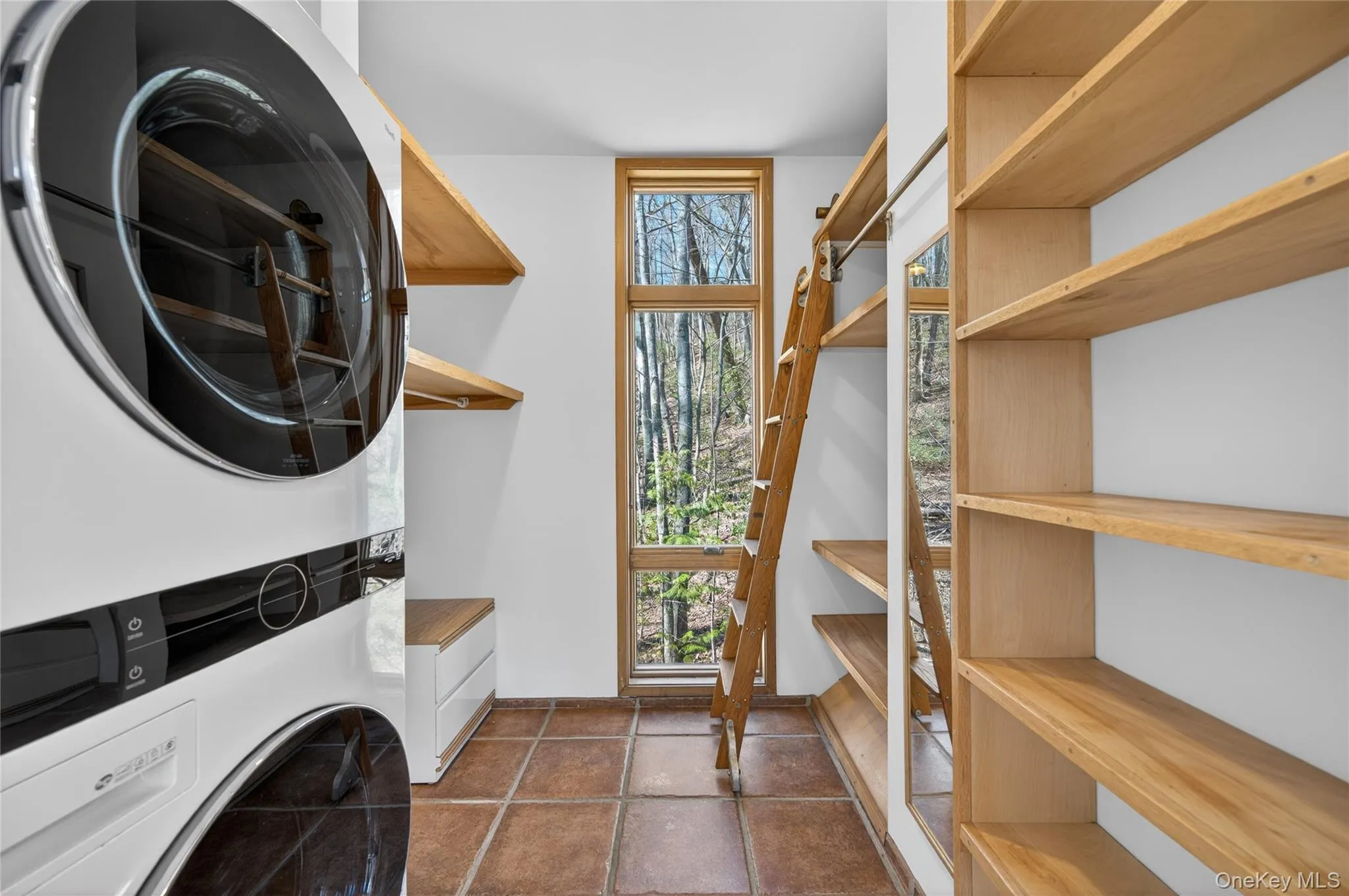 Laundry area featuring a stacked washer and dryer, extensive built-in shelving, a wood-frame window, and terracotta-style floor tiles Laundry area featuring a stacked washer and dryer, extensive built-in shelving, a wood-frame window, and terracotta-style floor tiles