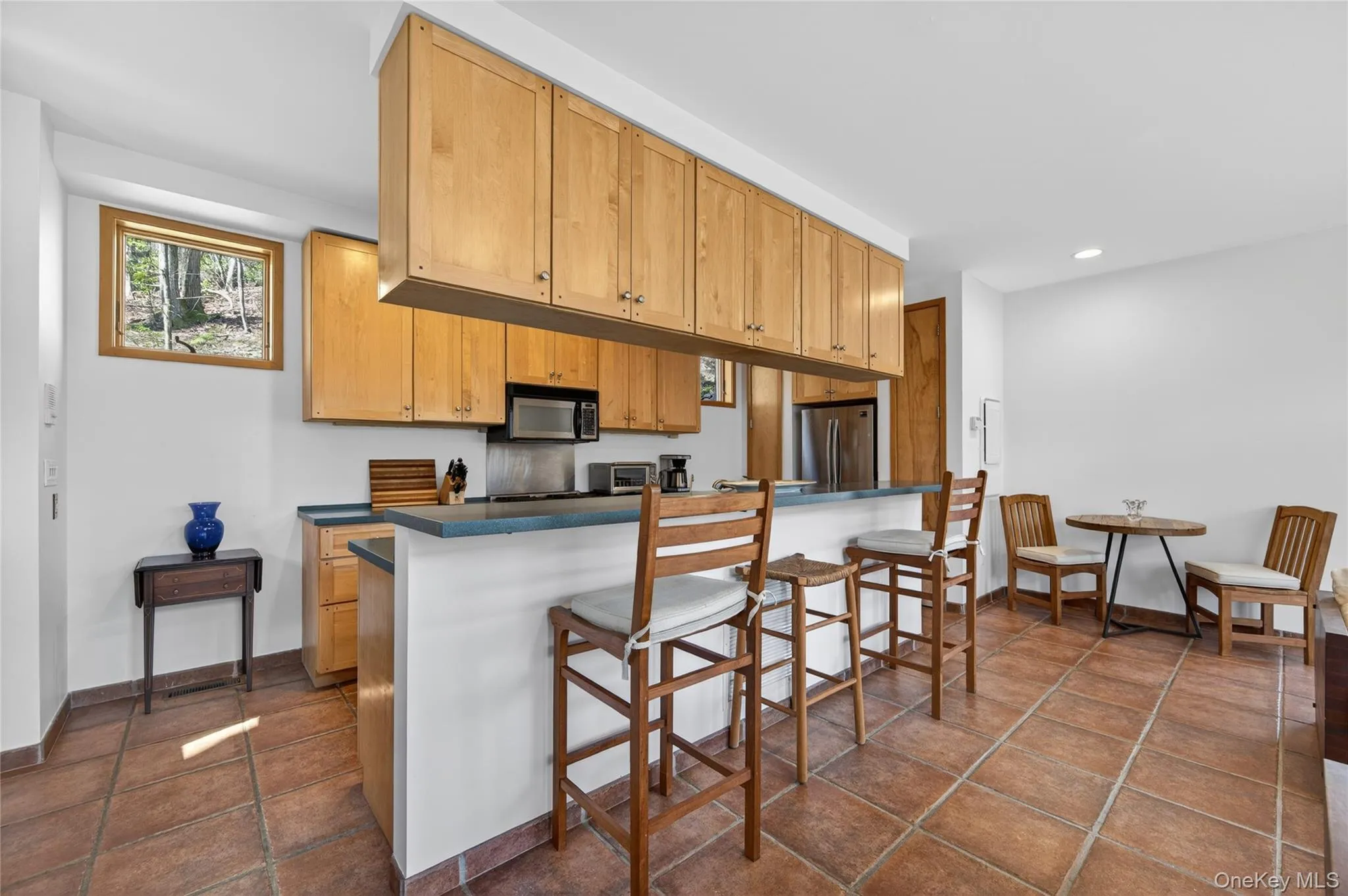 Kitchen featuring light wood cabinetry, stainless steel appliances, dark countertops, and terra cotta tile flooring Kitchen featuring light wood cabinetry, stainless steel appliances, dark countertops, and terra cotta tile flooring